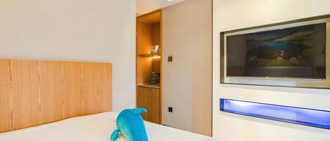 A modern hotel room with a wooden headboard, a large bed, a mounted TV, and a blue decorative object on the bed.