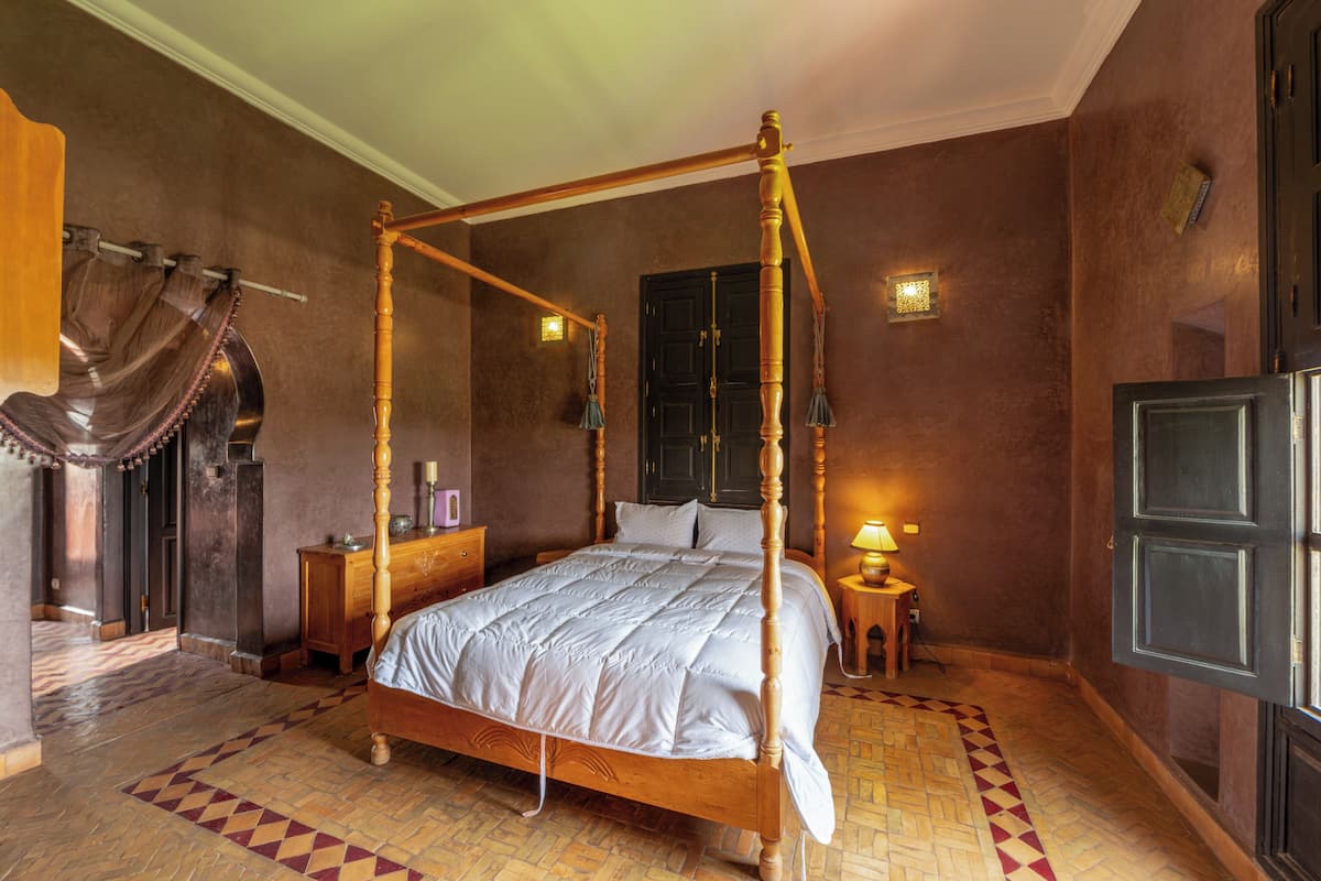 A four-poster bed in a room with a herringbone-patterned floor, a wooden nightstand, a lamp, and a dark door.