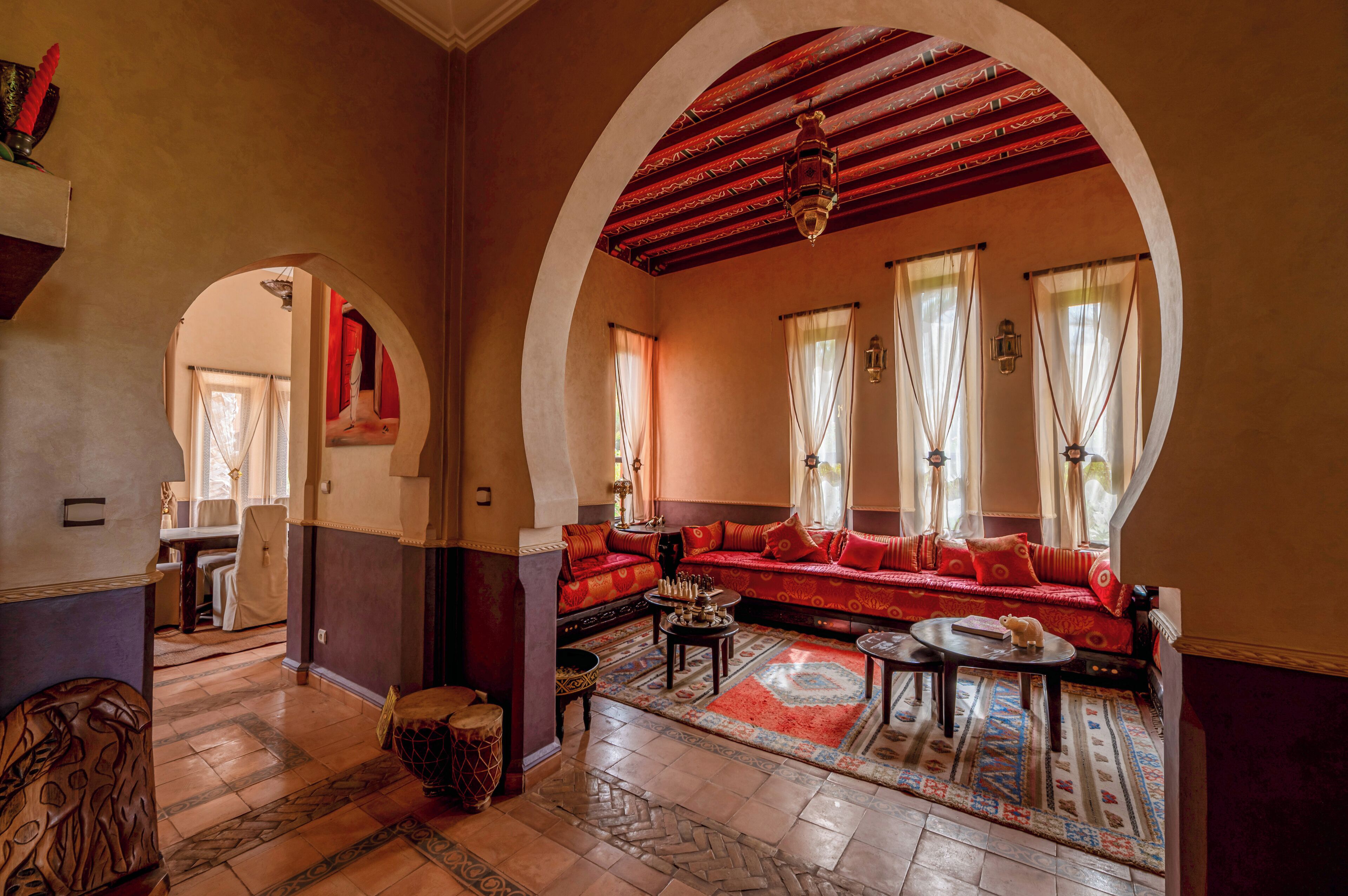 Un ampio soggiorno con un divano rosso, un tavolino da caffè e un tappeto decorato. Due aperture ad arco incorniciano la stanza, e sullo sfondo si intravede una zona pranzo.