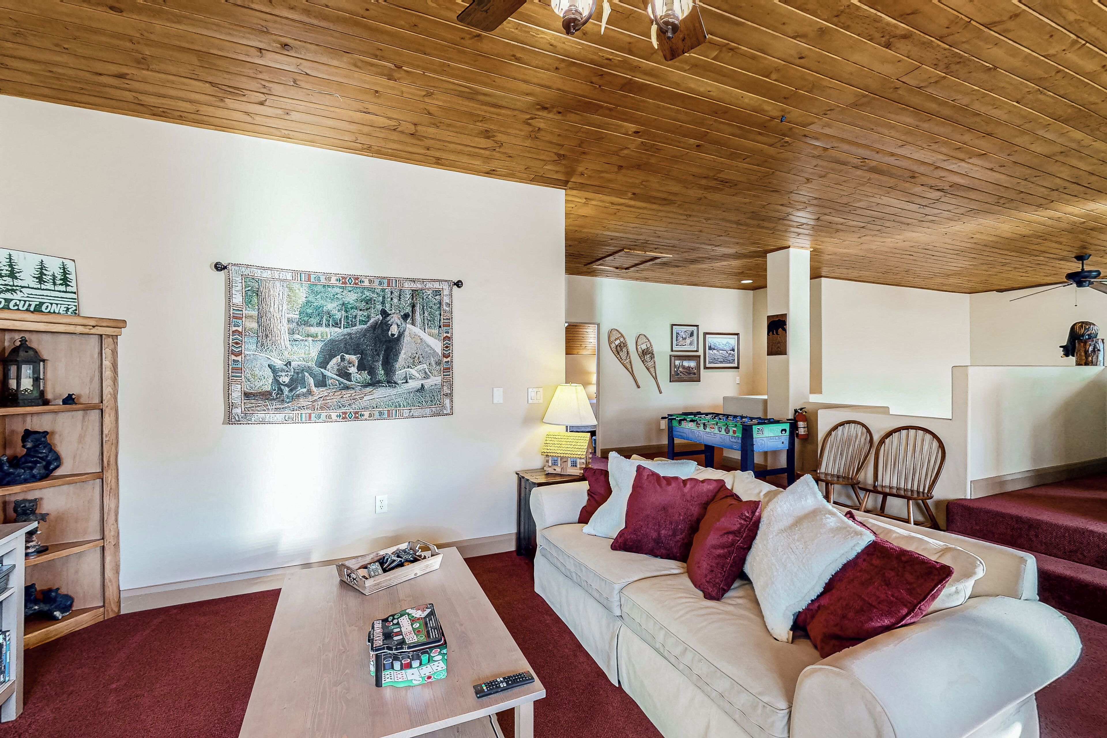 A cozy living room with a wooden ceiling, a beige sofa with red cushions, a wooden coffee table, and a mounted poster of a bear and a horse.