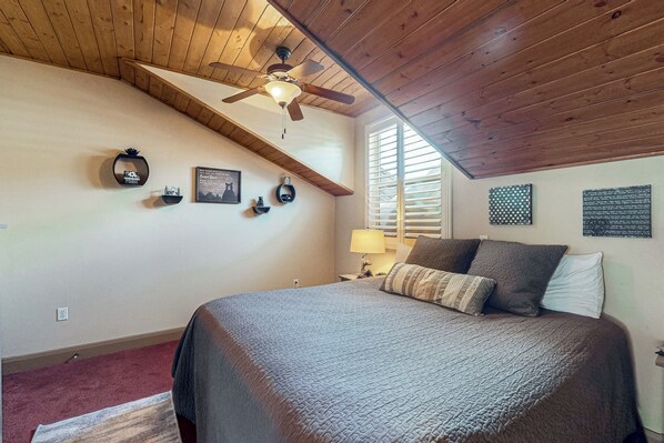 Ein Schlafzimmer mit schrägen Wänden, einem Bett mit dunkler Decke, einem Deckenventilator und Wanddekoration, darunter ein gerahmtes Bild und ein kleines Regal.