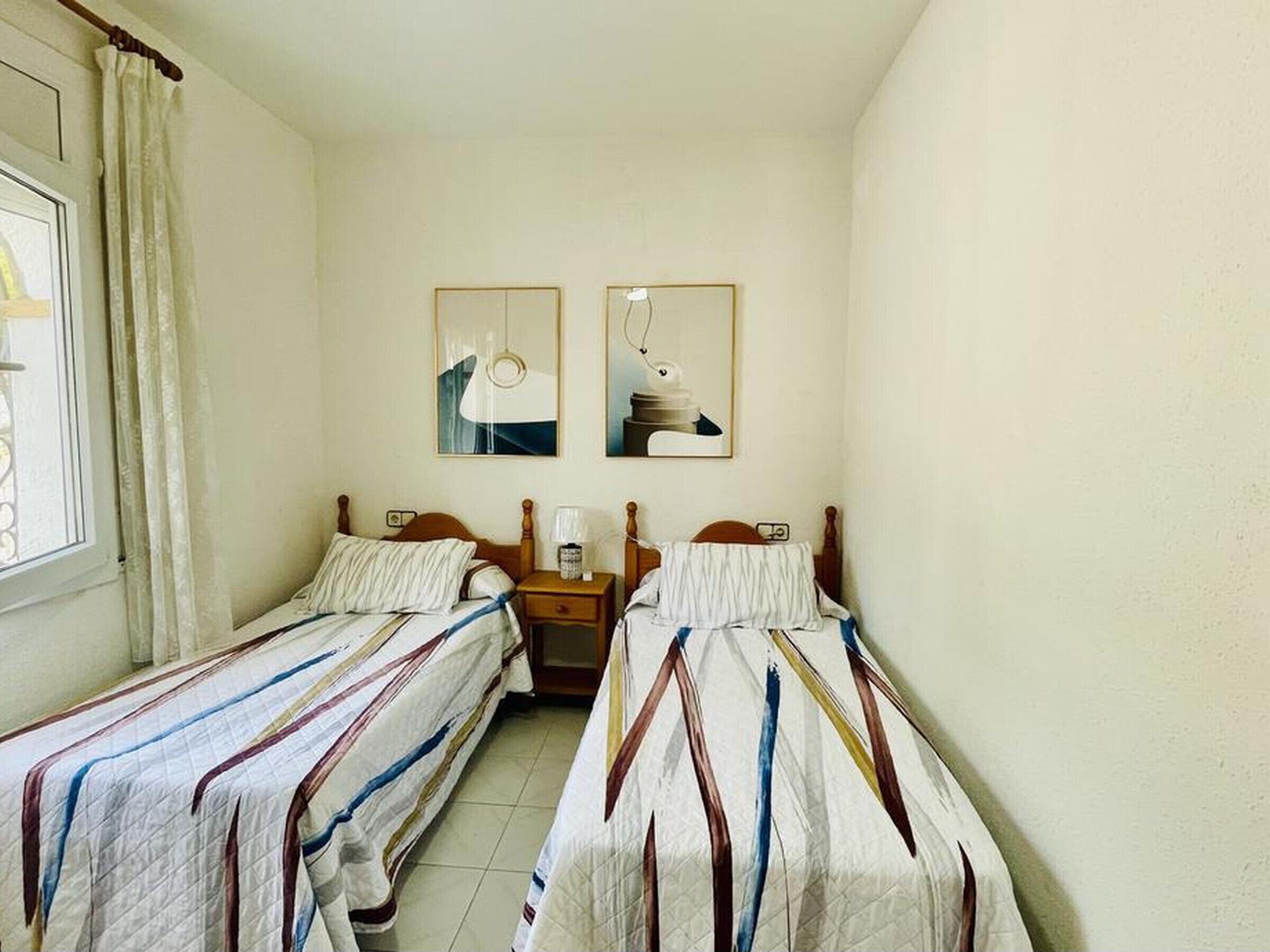 Een kleine, schone kamer met twee eenpersoonsbedden, een raam met gordijnen en twee foto's in lijsten aan de muur.