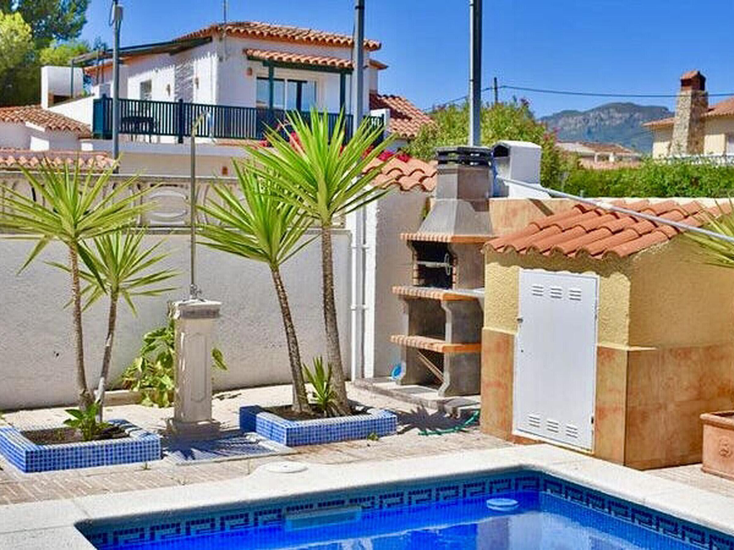 A swimming pool with a tiled edge, surrounded by palm trees and a building with a tiled roof.