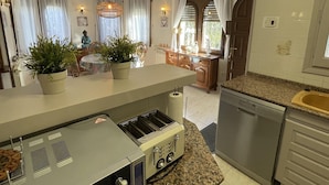 Cocina compacta con horno empotrado, pileta y mesada. Hay dos plantas en macetas sobre la mesada, y la cocina está conectada a un comedor con mesa y sillas.