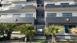 Un edificio moderno de varios pisos con balcones, una escalera central y un jardín bien cuidado con palmeras.