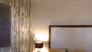 A bedroom with a wooden wall panel, a bedside table with a lamp, a bed with two pillows, and a window with blinds. - Beachfront Apartment with Garden, Pets Allowed (Miami Playa)