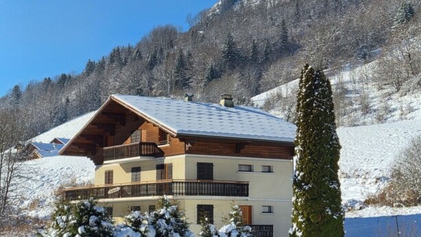 A multi-story chalet with a sloped roof, wooden balconies, and a view of a snowy mountain landscape. - Spacious 4-bedroom apartment with balcony and WIFI, near Châtel and La Chapelle (Abondance)
