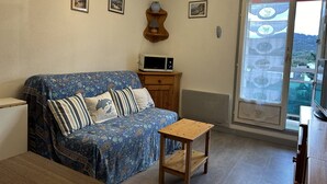 Een gezellige woonkamer met een gestreept bankstel, een klein houten tafeltje en een magnetron op een houten kast. Er hangen ingelijste foto's aan de muur en er is uitzicht naar buiten door een glazen deur.