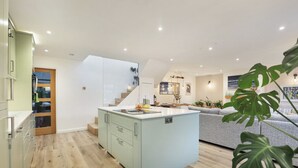 Una cocina moderna con una isla central, una escalera que conduce a un nivel superior y un comedor con sofá y plantas.