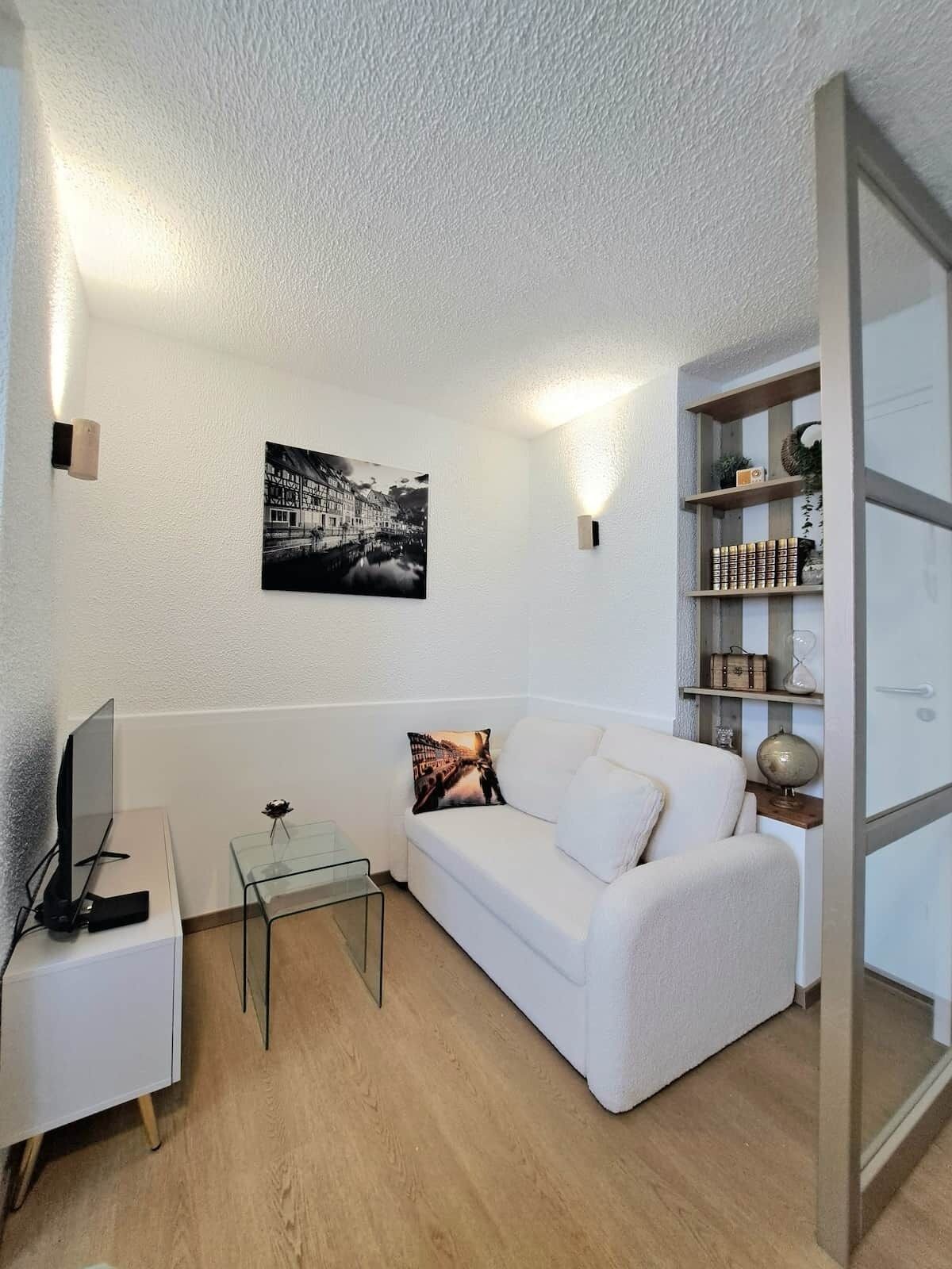Ein Wohnzimmer mit einem weißen Sofa, einem Glastisch, einem Flachbildfernseher und einem Bücherregal mit Dekoration.