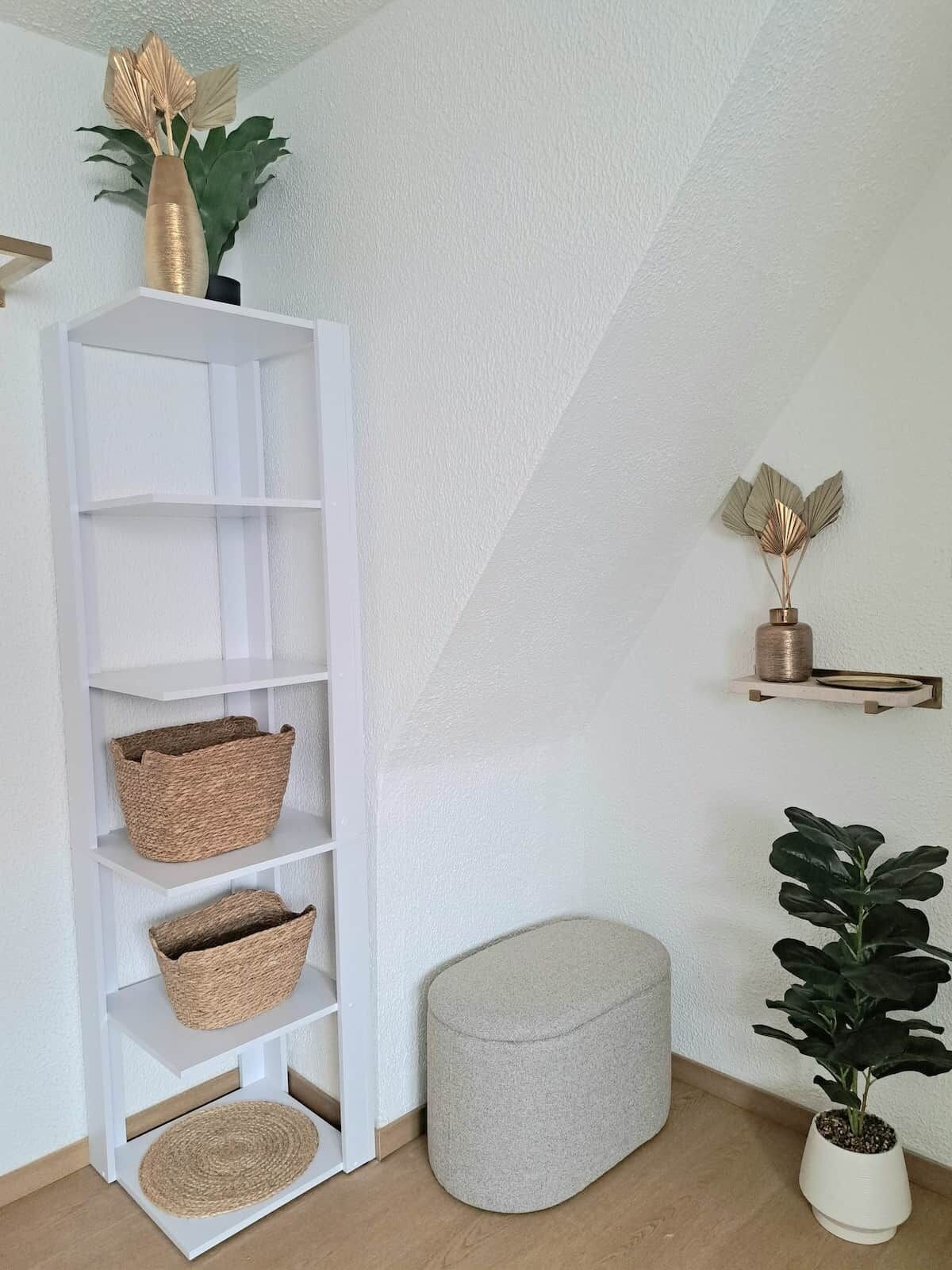 Un estante blanco con cestas tejidas, un otomano gris, una planta en maceta y objetos decorativos sobre una pared blanca.