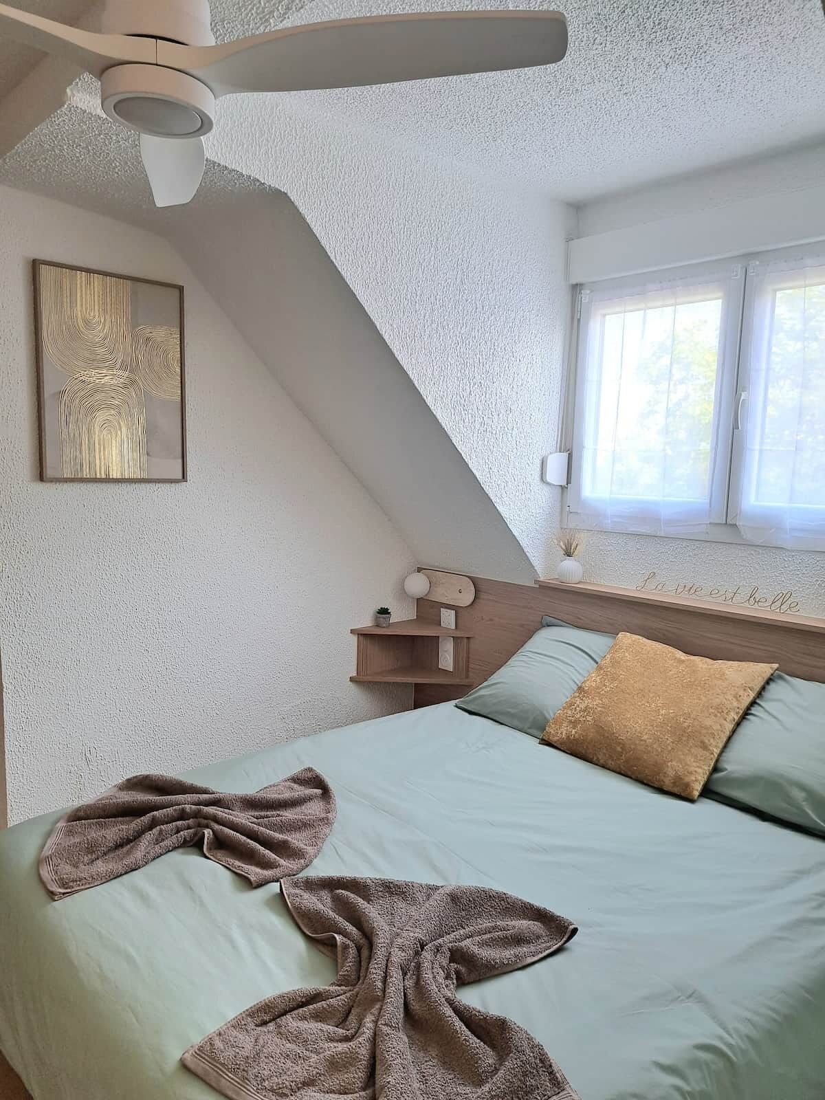 A bedroom with a bed, a ceiling fan, a window, and a wall-mounted shelf.