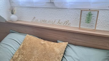 A bed with a wooden headboard, a decorative sign, a framed plant, and a vase with dried flowers.