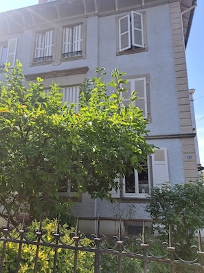 A multi-story building with white shutters on windows, a visible number '25' on the side, and a metal fence in the foreground. - To complete (Colmar)
