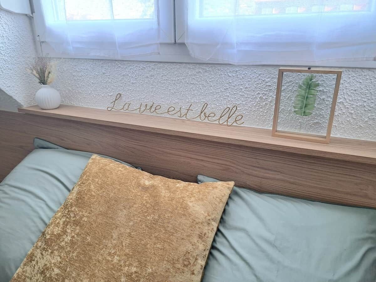 Ein Bett mit Holz-Kopfstück, ein dekoratives Schild, eine gerahmte Pflanze und eine Vase mit getrockneten Blumen.