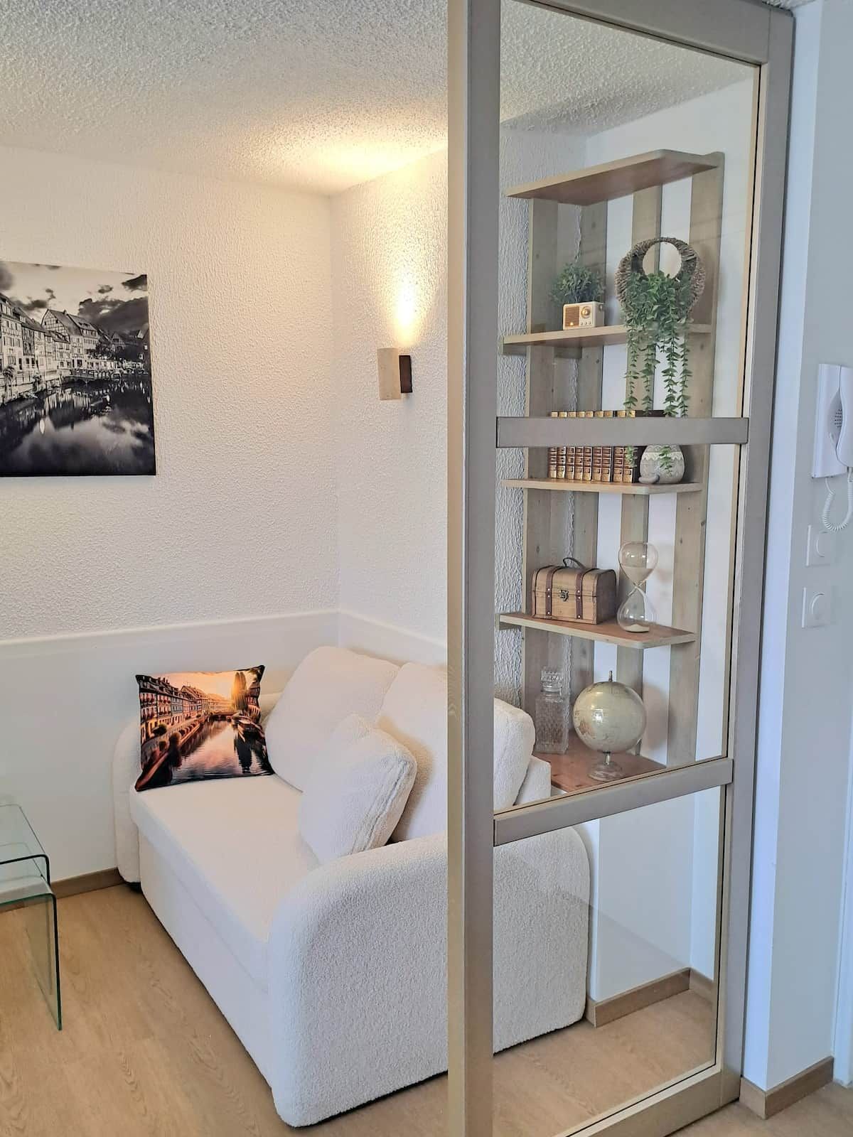 Una sala moderna con un sofá blanco, una mesa de centro de vidrio, un cojín decorativo, un cuadro en la pared y un estante con puerta de vidrio que contiene diversos objetos.
