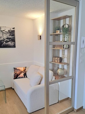 A modern living room with a white sofa, a glass coffee table, a decorative pillow, a wall-mounted picture, and a glass-fronted shelving unit with various items. - To complete (Colmar)