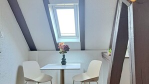 A small, modern room with a sloped ceiling, a skylight, a round table with two chairs, and string lights. - To complete (Colmar)