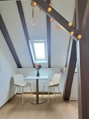 A small, modern room with a sloped ceiling, a skylight, a round table with two chairs, and string lights. - To complete (Colmar)