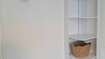 A white wall with a gold metal rack holding a decorative item, a white shelving unit with baskets, and a small woven basket on the floor.