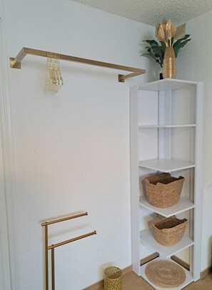 A white wall with a gold metal rack holding a decorative item, a white shelving unit with baskets, and a small woven basket on the floor. - To complete (Colmar)