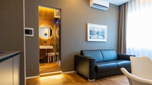 A modern hotel room with a wooden floor, a black leather sofa, a white armchair, a framed picture on the wall, and a glimpse of a bathroom with a mirror and sink. - B&Z Luxury (Bari)