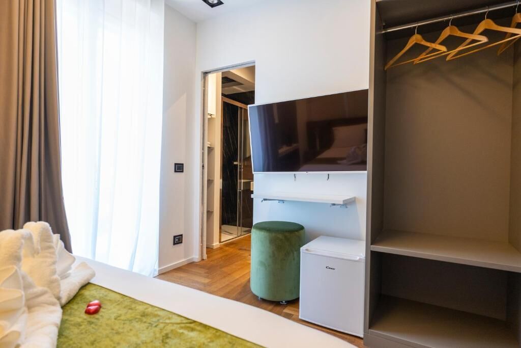 A modern hotel room with a large wardrobe, a flat-screen TV, a small fridge, and a green ottoman.