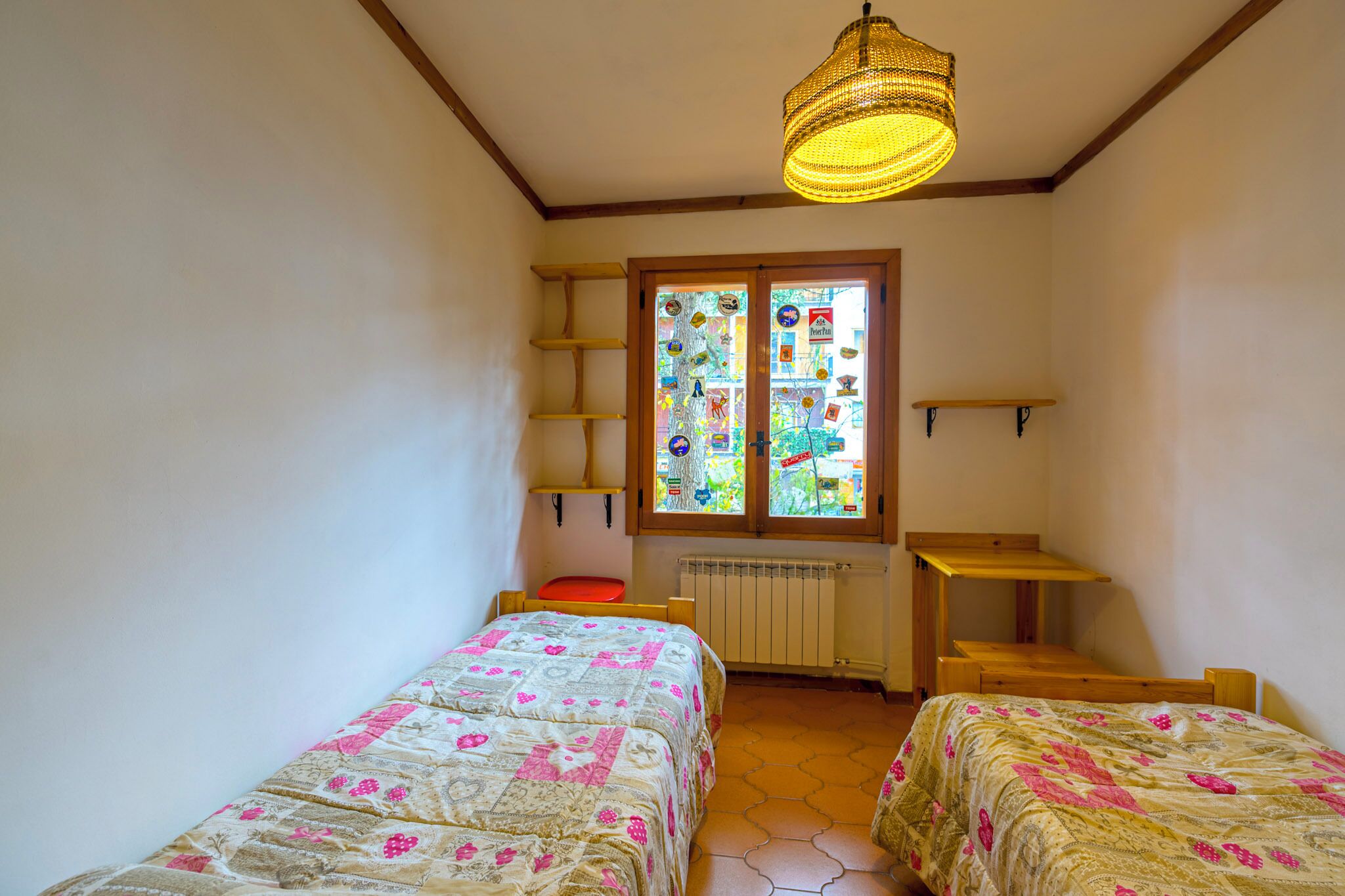 A small room with two beds, a window with colorful decorations, a wooden shelf, and a hanging light fixture.