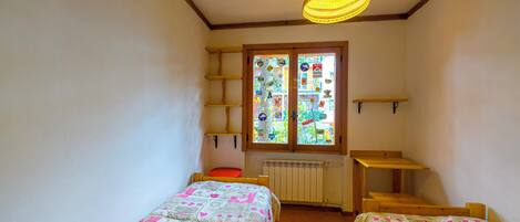 A small room with two beds, a window with colorful decorations, a wooden shelf, and a hanging light fixture.