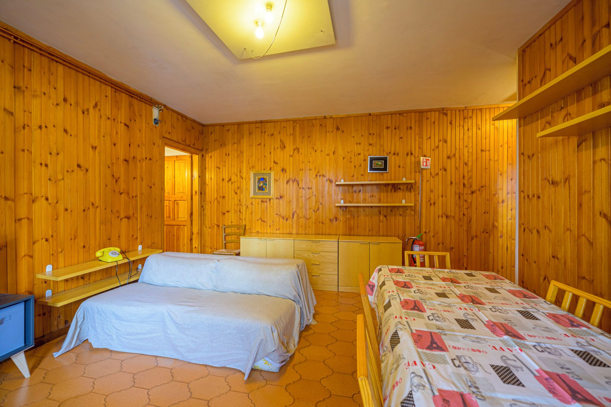 Una habitación con paredes de madera y una lámpara en el techo. Hay dos camas, un escritorio y una silla.