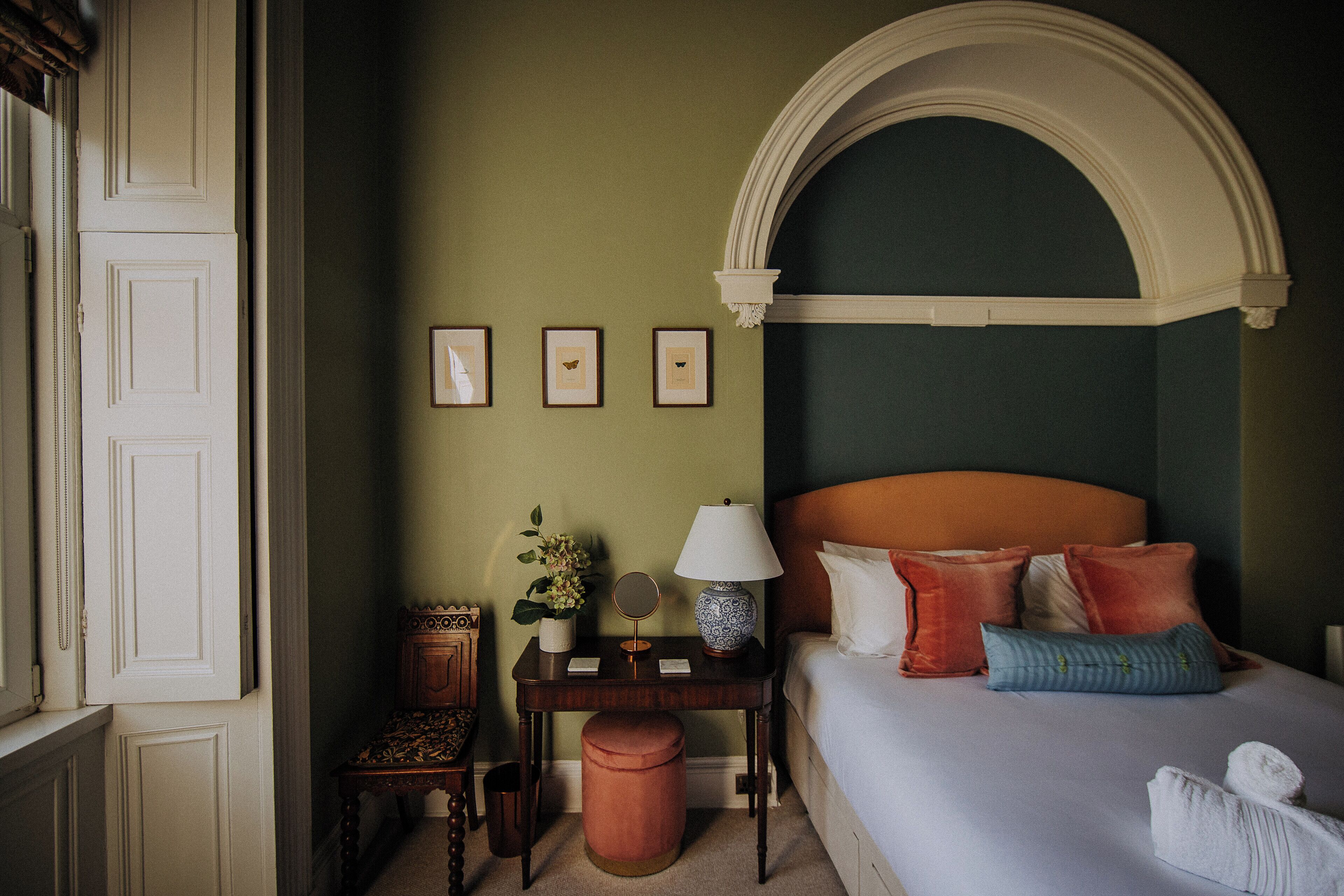 Une chambre d’hôtel avec un lit, une table de chevet, une lampe, un miroir et des tableaux encadrés accrochés au mur.