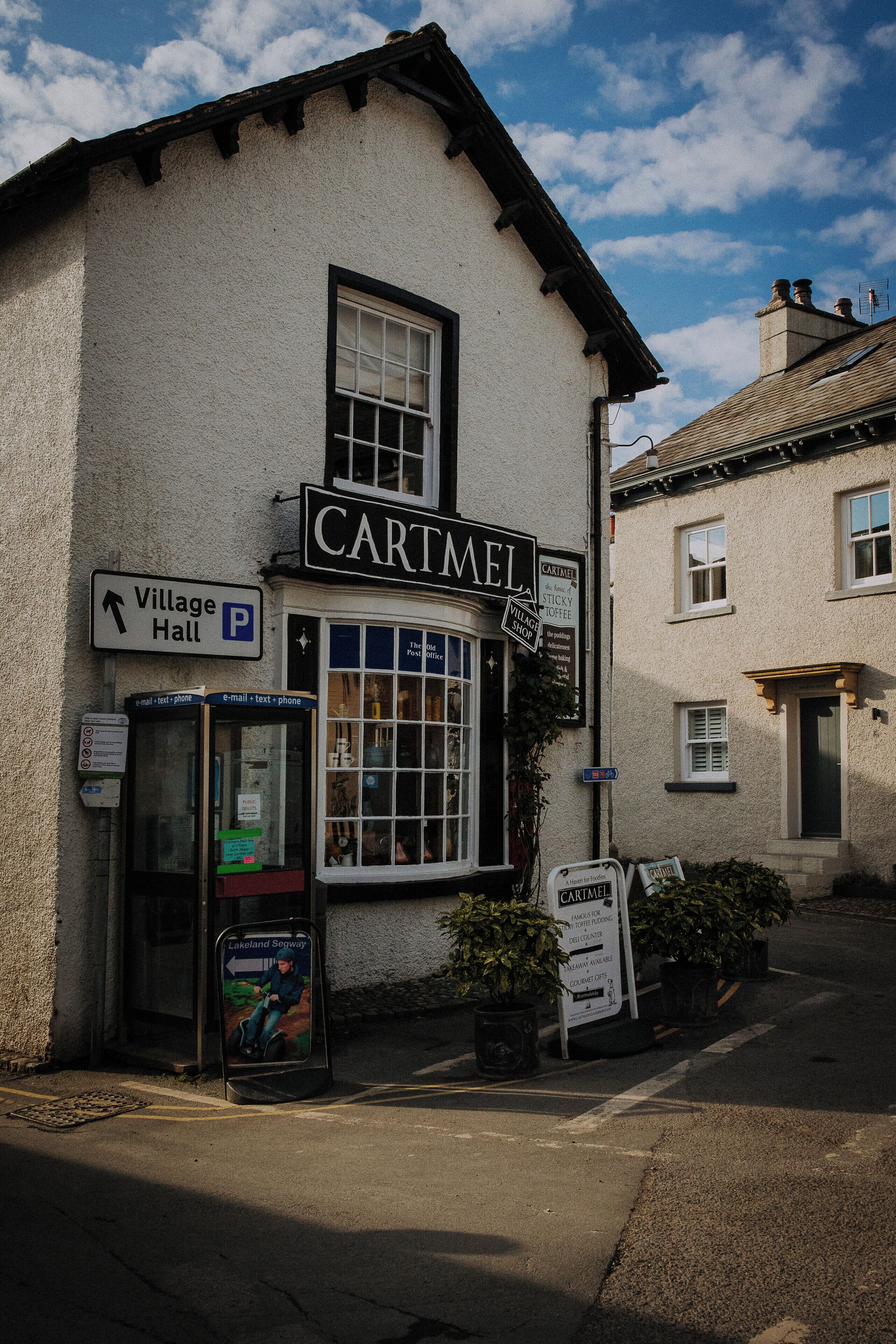 A building with a sign reading "CARTMEL" and a Village Hall sign pointing to the left.