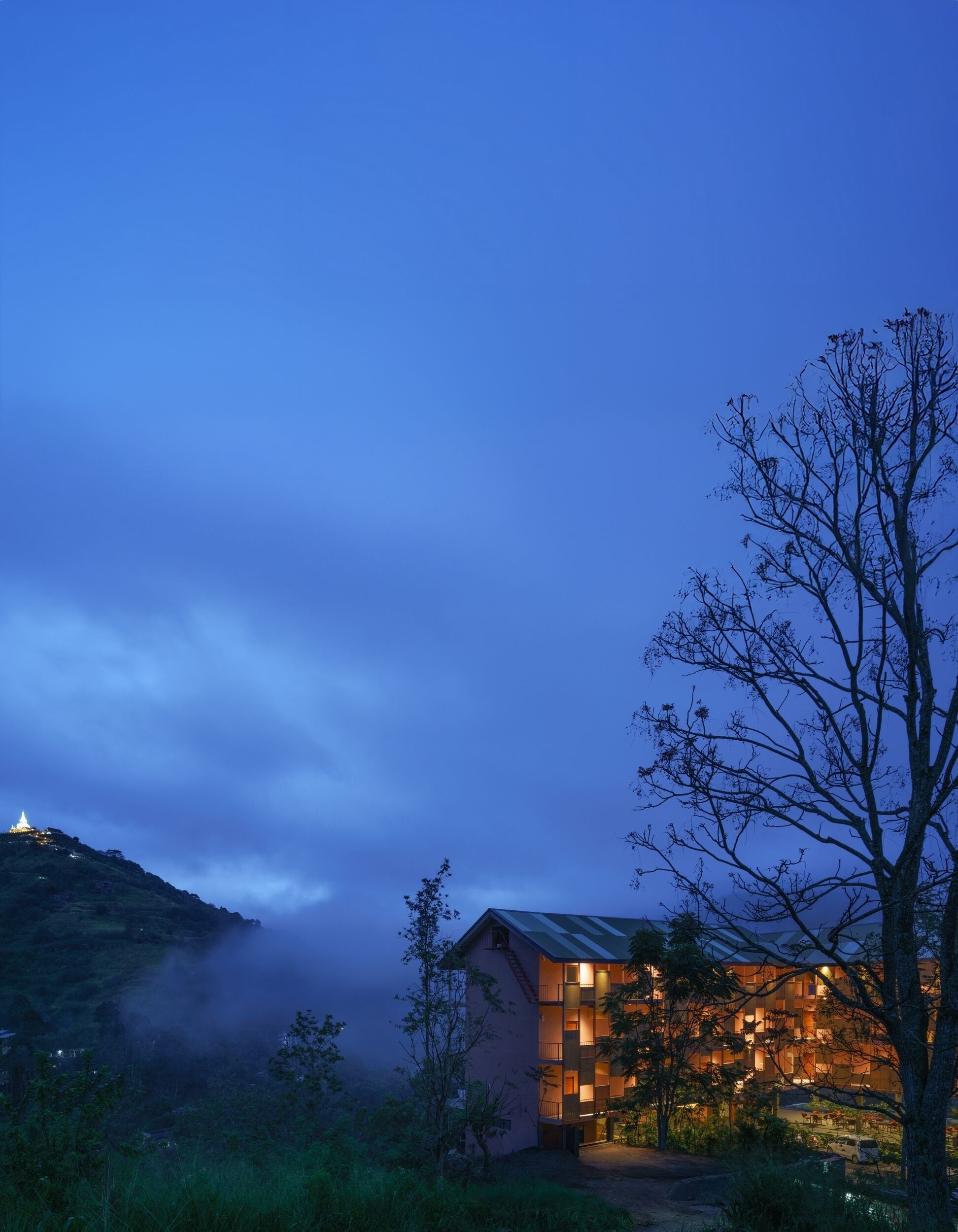 A building with multiple floors and balconies, surrounded by trees and a hill in the background.