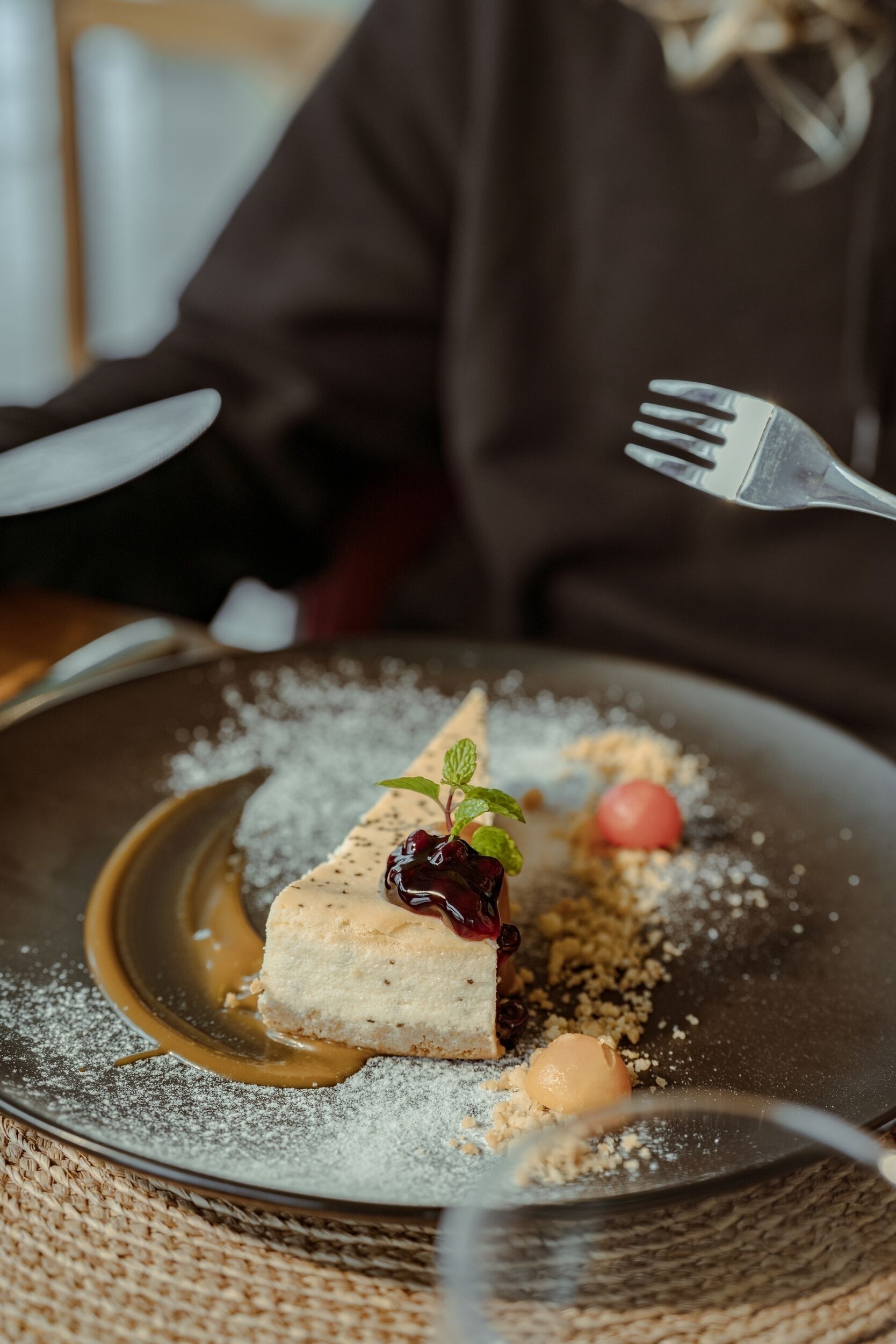 A dessert plate with a slice of cheesecake, a cherry, and a mint leaf on a dark plate.