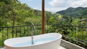 Una bañera blanca independiente en un balcón con baranda de metal, con vistas a un paisaje verde exuberante y una montaña al fondo.