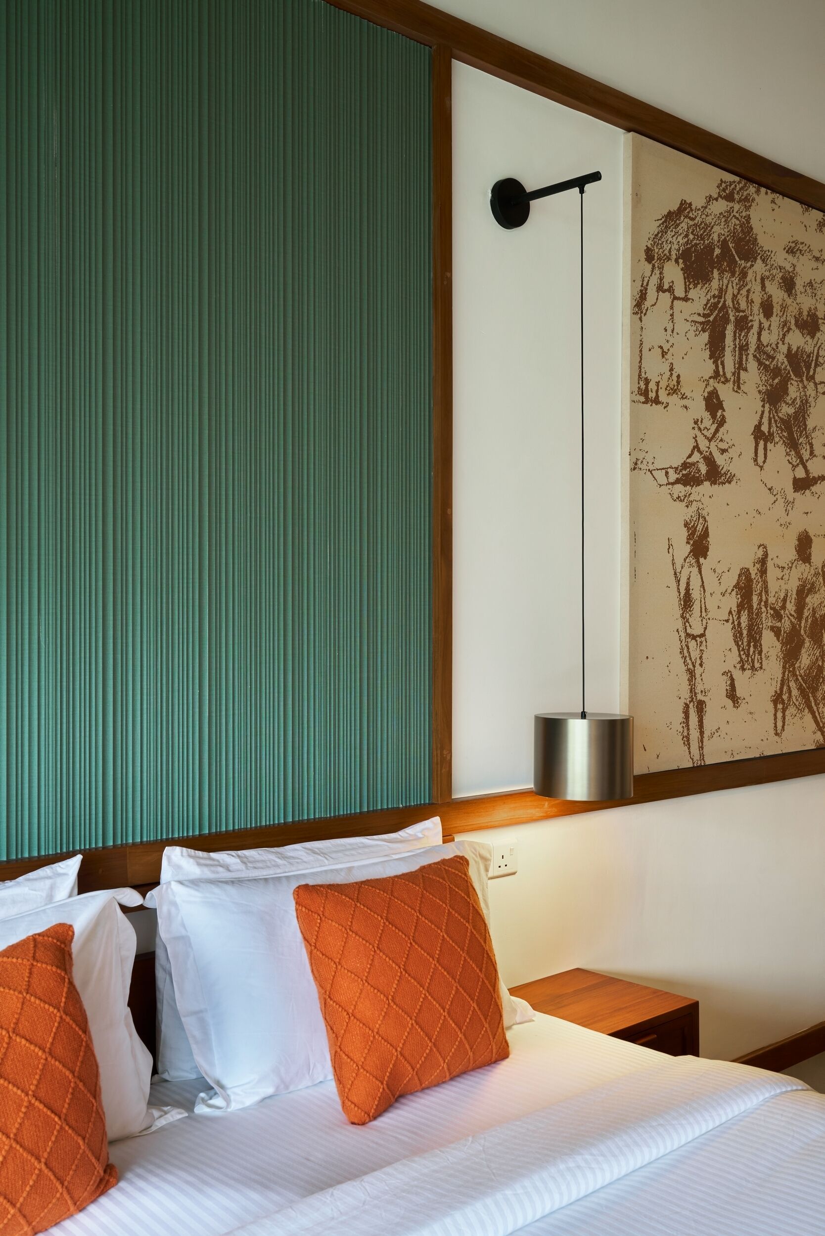 A hotel room with a bed, two orange pillows, a nightstand, a wall-mounted lamp, and a textured artwork.