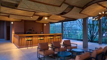 Un área de bar moderna con barra, asientos y ventanas amplias que ofrecen vistas a árboles y edificios.