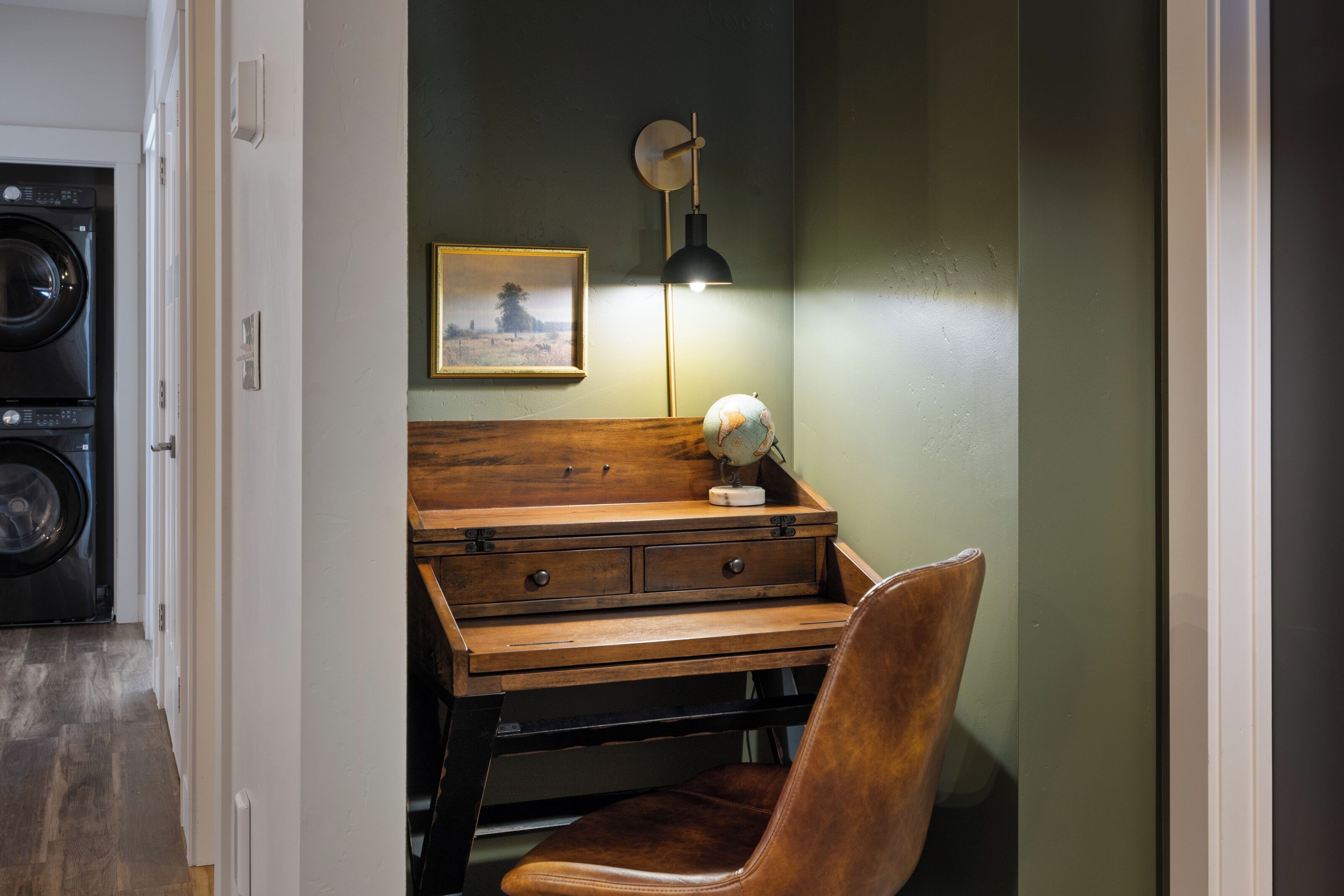 A room with a wooden desk, a chair, a wall-mounted lamp, and a framed picture.