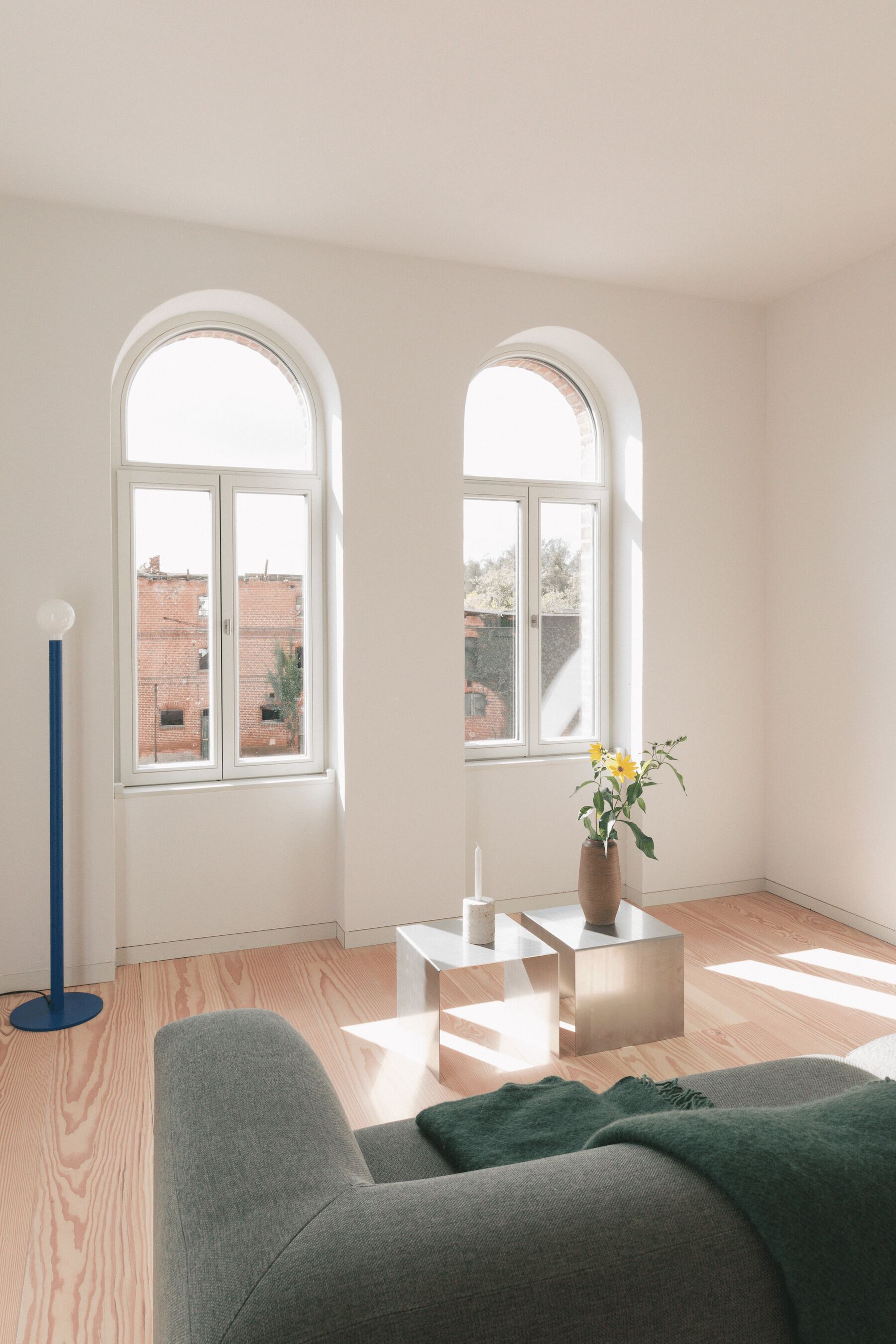 Una habitación con dos ventanas arqueadas, un sofá verde, una pequeña mesa con un jarrón de flores y una lámpara de pie azul alta.