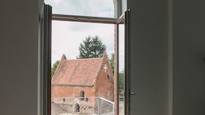 A room with a wooden floor, a grey sofa, and a large arched window leading to an outdoor area with a brick building and a fence.