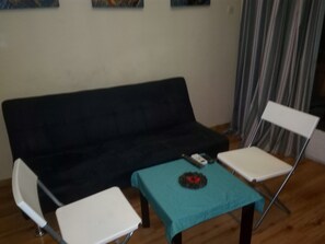 Ein Wohnbereich mit schwarzem Sofa, weißem Sessel, kleinem Tisch mit türkisblauem Tischläufer und Fernbedienung auf dem Tisch.