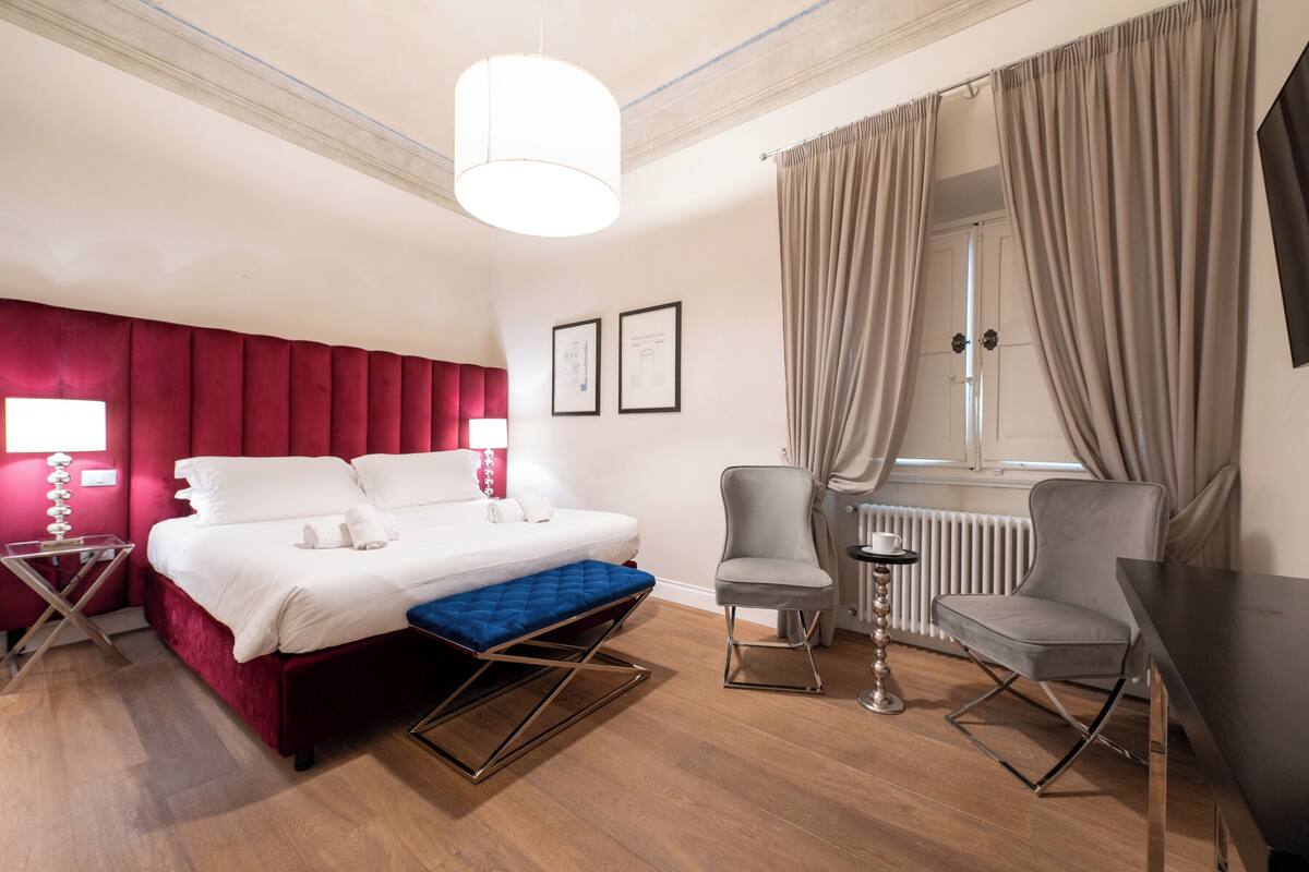 A hotel room with a bed, a desk, a chair, a radiator, and a window with curtains.