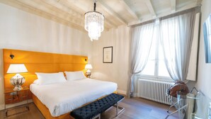 Une chambre d’hôtel avec une tête de lit en bois, un grand lit avec une literie blanche, un banc au pied du lit, une table en verre avec une chaise, un lustre et une fenêtre avec des rideaux.
