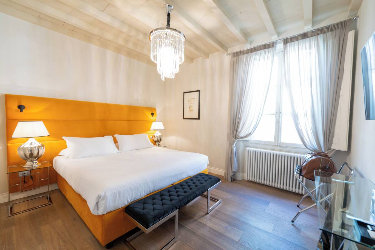 A hotel room with a wooden headboard, a large bed with white bedding, a bench at the foot of the bed, a glass table with a chair, a chandelier, and a window with curtains.