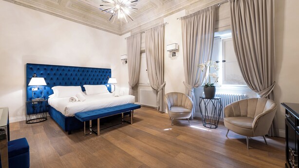 Une chambre d’hôtel moderne avec un lit doté d’une tête de lit bleue à motifs capitonnés, un banc, une chaise et une petite table sur laquelle est posée une plante en pot.