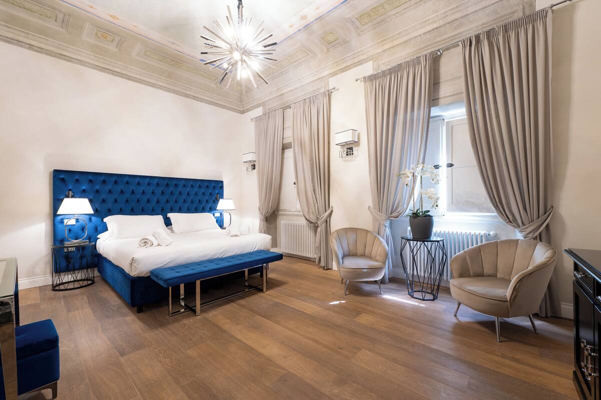 A modern hotel room with a blue tufted headboard bed, a bench, a chair, and a small table with a potted plant.