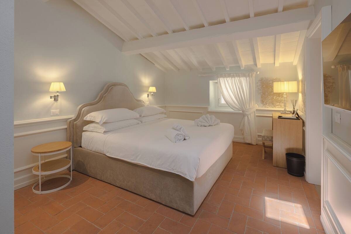 A neatly arranged bedroom with a large bed, two bedside lamps, a small round table, and a window with curtains.