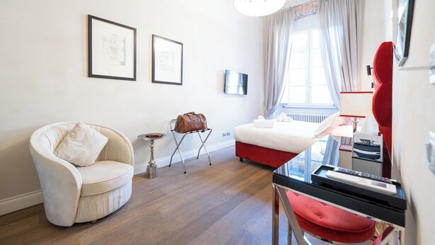 Una habitación de hotel moderna con un sillón blanco curvo, un sofá rojo, una mesita auxiliar pequeña y una cama con cabecera roja.