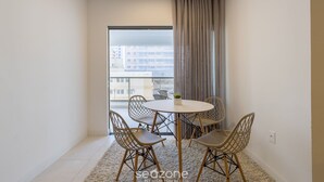 Une petite pièce moderne, dotée d’une table ronde et de quatre chaises, d’une grande fenêtre offrant une vue sur les immeubles et d’un rideau à la fenêtre.