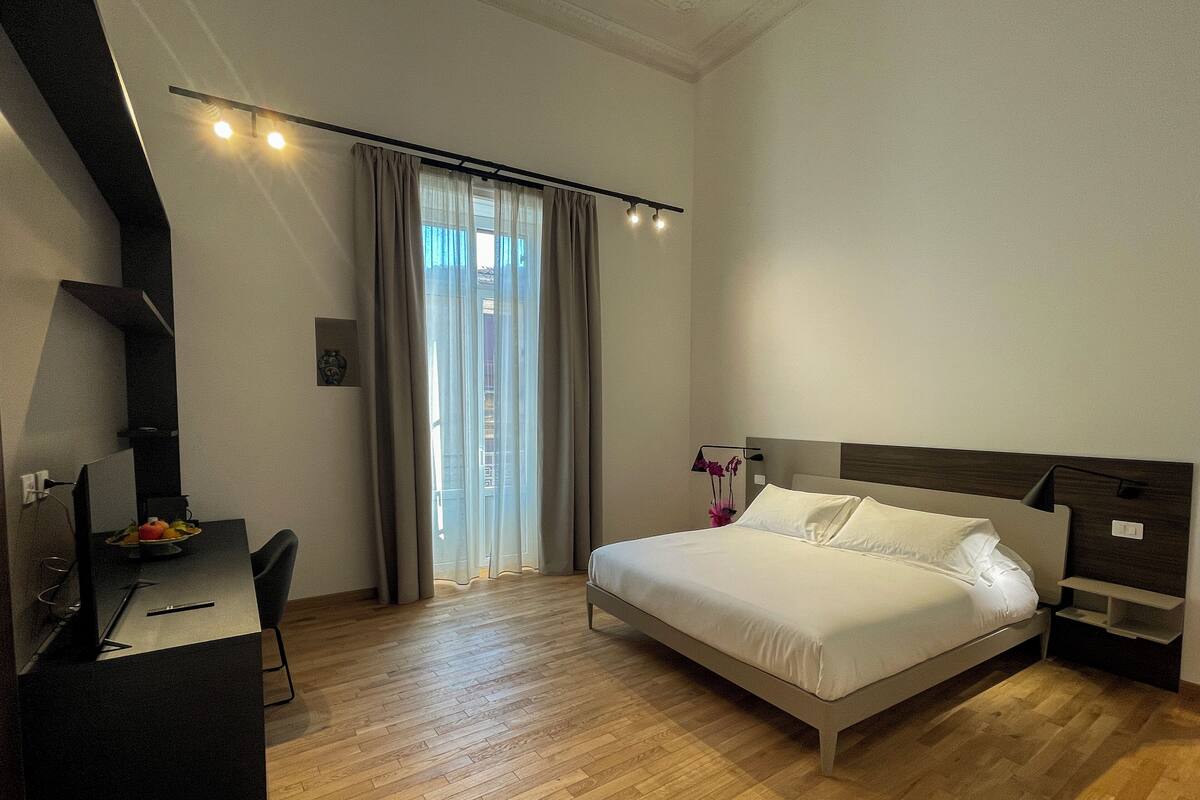 Una camera d'albergo moderna con pavimento in legno, un letto con lenzuola bianche, una scrivania con un computer e una finestra con tende.